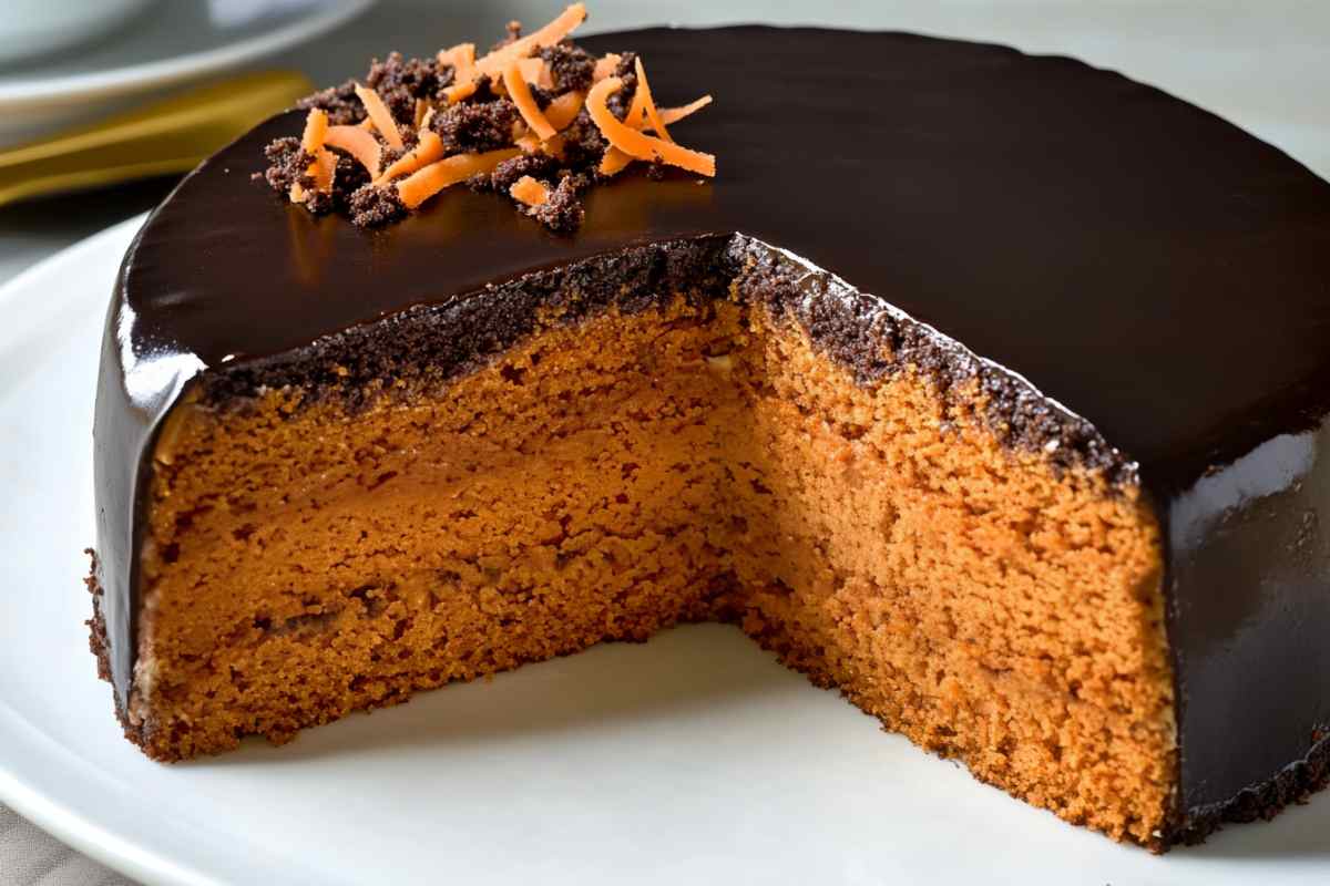 A slice of Brazilian carrot cake with a rich chocolate glaze on top, garnished with shredded carrots and chocolate crumbs, showing its moist orange interior on a white plate