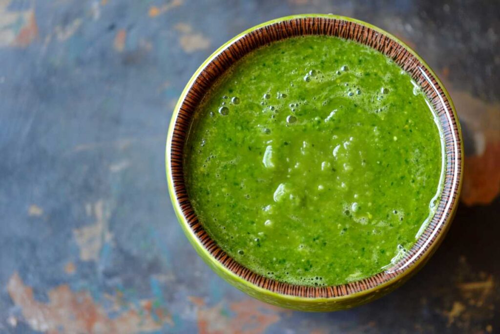 A bowl of vibrant green gazpacho, smooth and garnished with small bubbles, set against a rustic background