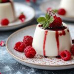 A serving of panna cotta drizzled with raspberry sauce, garnished with fresh raspberries and a mint leaf, on a decorative plate