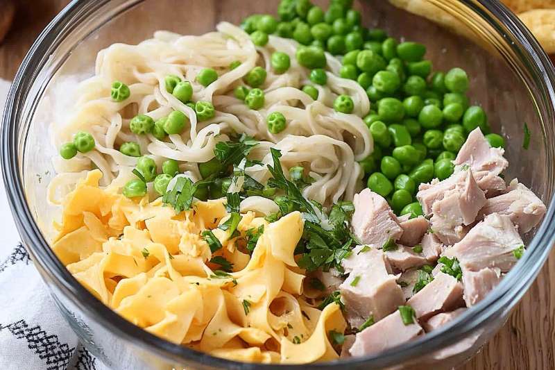 bowl filled with ingredients for a tuna casserole, including pasta, green peas, diced tuna, and chopped parsley.